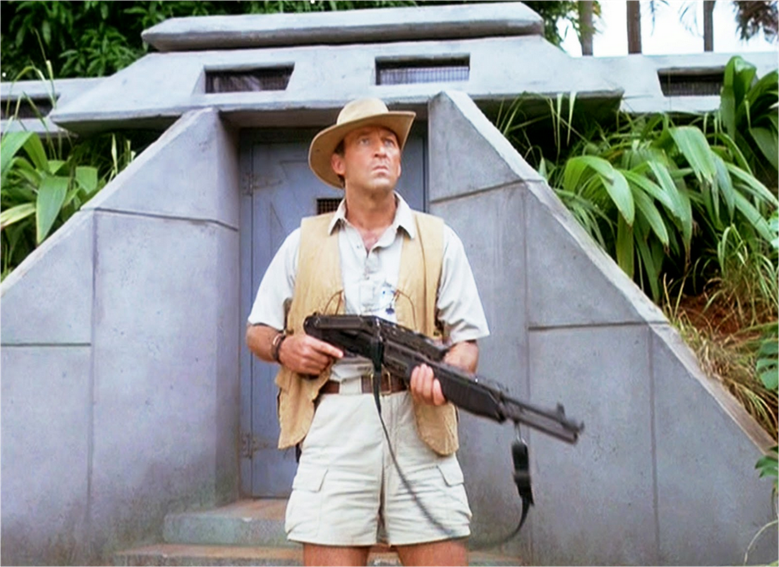 A photo of Muldoon, Hammond's game warden in front of a buttressed side entrance to the visitors' center. Muldoon is kitted out in khaki safari gear and sports a rifle.