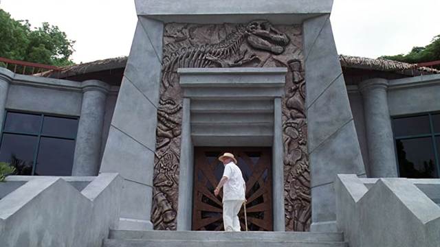 A shot of the entrance to the visitors' center from the landing of the entryway steps. Hammond is framed in the doorway. He is wearing a white linen safari suit.
